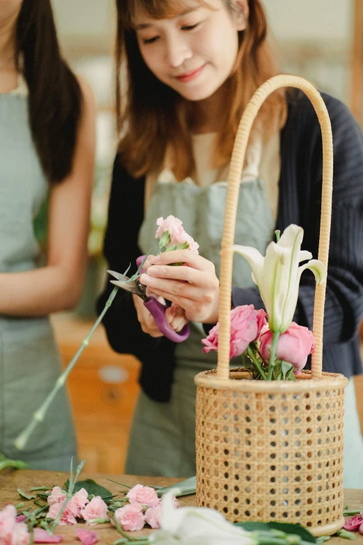 a woman standing in front of a basket filled with flowers, trending on pexels, in a workshop, holding a rose in a hand, staff, bustling