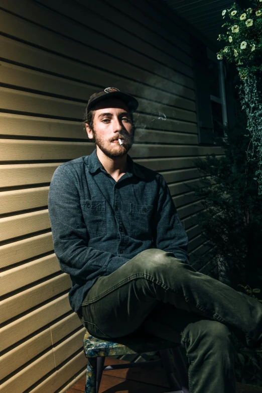 a man sitting on a chair in front of a building, a portrait, by Andrew Stevovich, unsplash, mac miller, smoky lighting, cigarette in his mouth, profile image