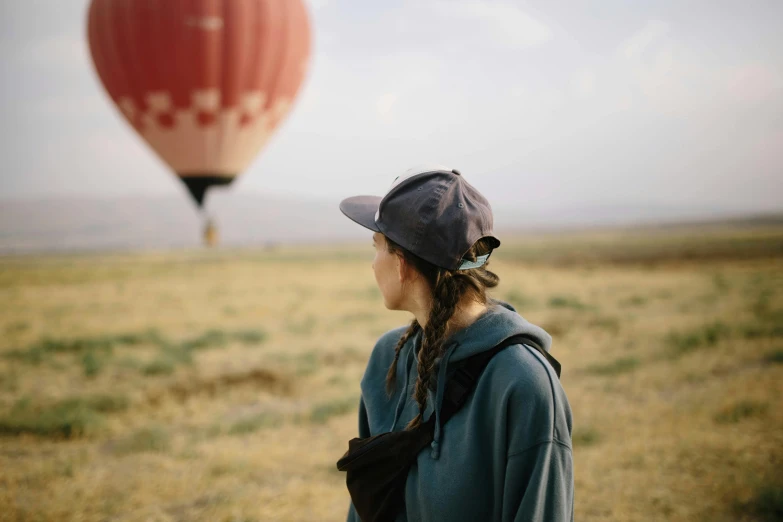 a woman standing in a field looking at a hot air balloon, wearing a red backwards cap, lachlan bailey, looking off into the distance, high elevation