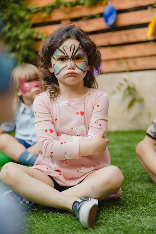 a group of children sitting on top of a lush green field, face paint around eyes, frustrated face, insect trainer girl, at a birthday party