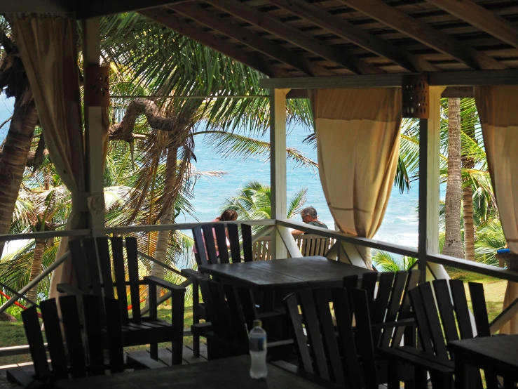 a view of the ocean from a covered porch, by Sam Dillemans, restaurant in background, chief keef in the garden of eden, cafe tables, rum