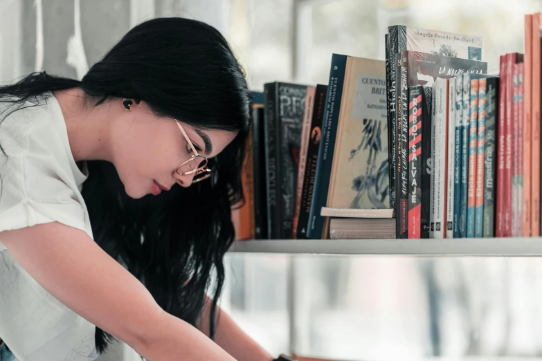 a woman reading a book in front of a bookshelf, pexels contest winner, wavy long black hair and glasses, profile image, thumbnail, bent over posture
