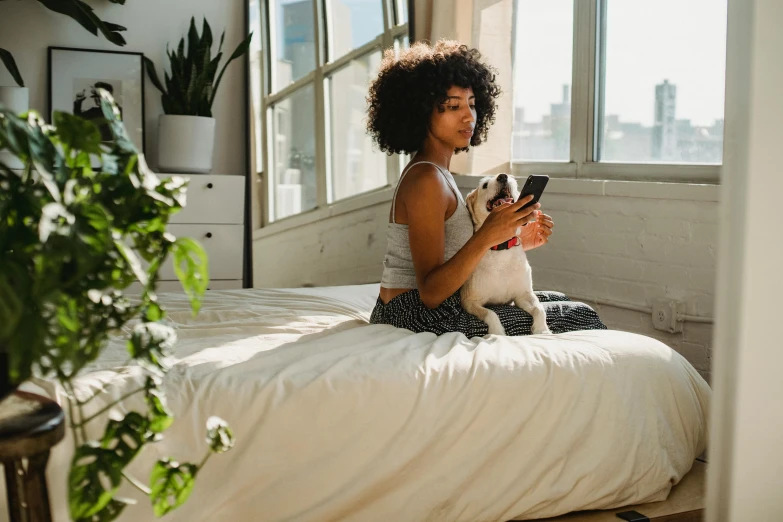 a woman sitting on a bed with a dog looking at her phone, trending on pexels, beautiful city black woman only, springtime morning, small dog, white bed