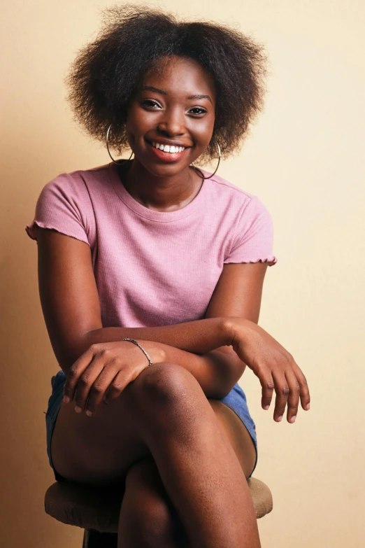 a woman sitting on top of a wooden chair, by Lily Delissa Joseph, pexels contest winner, photorealism, dark short curly hair smiling, black teenage girl, tan skin a tee shirt and shorts, plain background