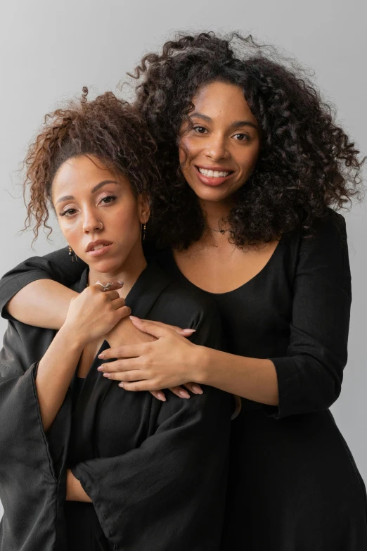 a couple of women standing next to each other, natural hair, wearing a black robe, on grey background, promo image