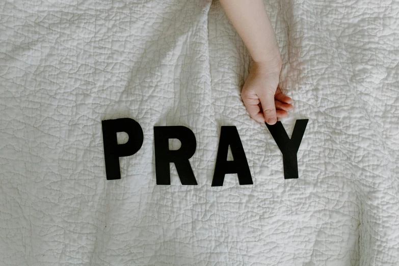 a person laying in bed with a white blanket with the word pray written on it, pexels, little boy wearing nun outfit, on grey background, back of hand on the table, softplay