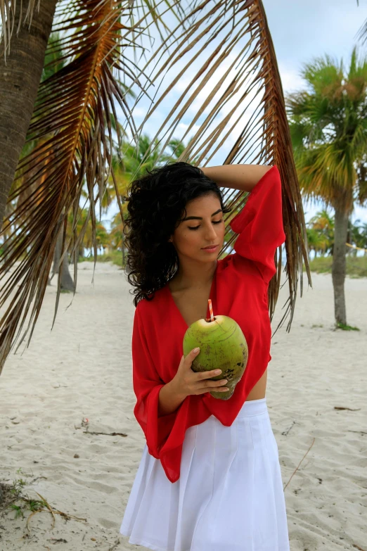 a woman standing on a beach holding a coconut, inspired by Ruth Orkin, unsplash, fine art, red shirt, imaan hammam, miami beach, modeled