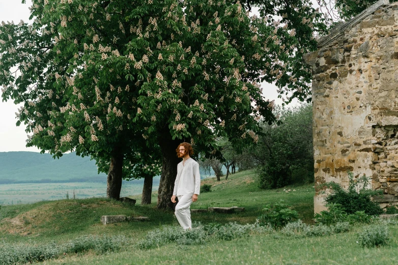 a man standing in a field next to a tree, inspired by Henri-Julien Dumont, pexels contest winner, renaissance, wearing white pajamas, ancient garden behind her, tuscany hills, standing outside a house