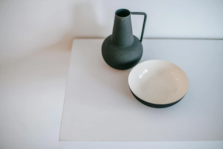 a black vase sitting on top of a table next to a white bowl, inspired by Constantin Hansen, unsplash, minimalism, zinc white, collection product, detail shot, plates