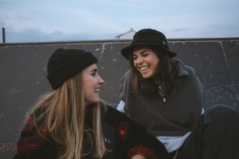 a couple of women sitting on top of a skateboard ramp, by Emma Andijewska, trending on pexels, beanie, smiling at each other, 15081959 21121991 01012000 4k, black pointed hat