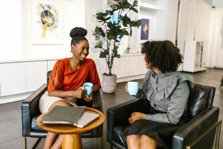 two women sitting at a table with a cup of coffee, pexels contest winner, happening, npc talking, sitting in a waiting room, both laughing, in an office