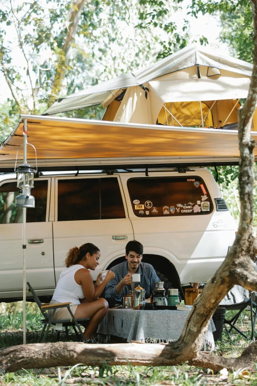 a couple of people sitting at a table under a tent, with a roof rack, jungle setting, coffee, van