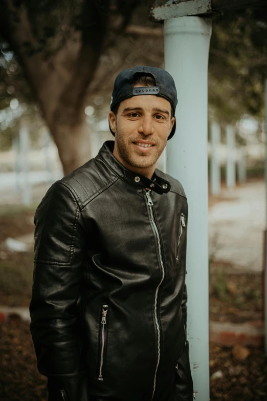 a man in a leather jacket standing next to a pole, inspired by Dicky Doyle, pexels contest winner, wearing baseball cap, smiling male, zachary corzine, frontal pose