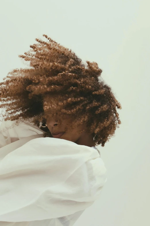 a woman with curly hair standing in front of a white wall, by Lily Delissa Joseph, trending on unsplash, hugging and cradling, his hair moves with the wind, muted browns, wearing a white robe