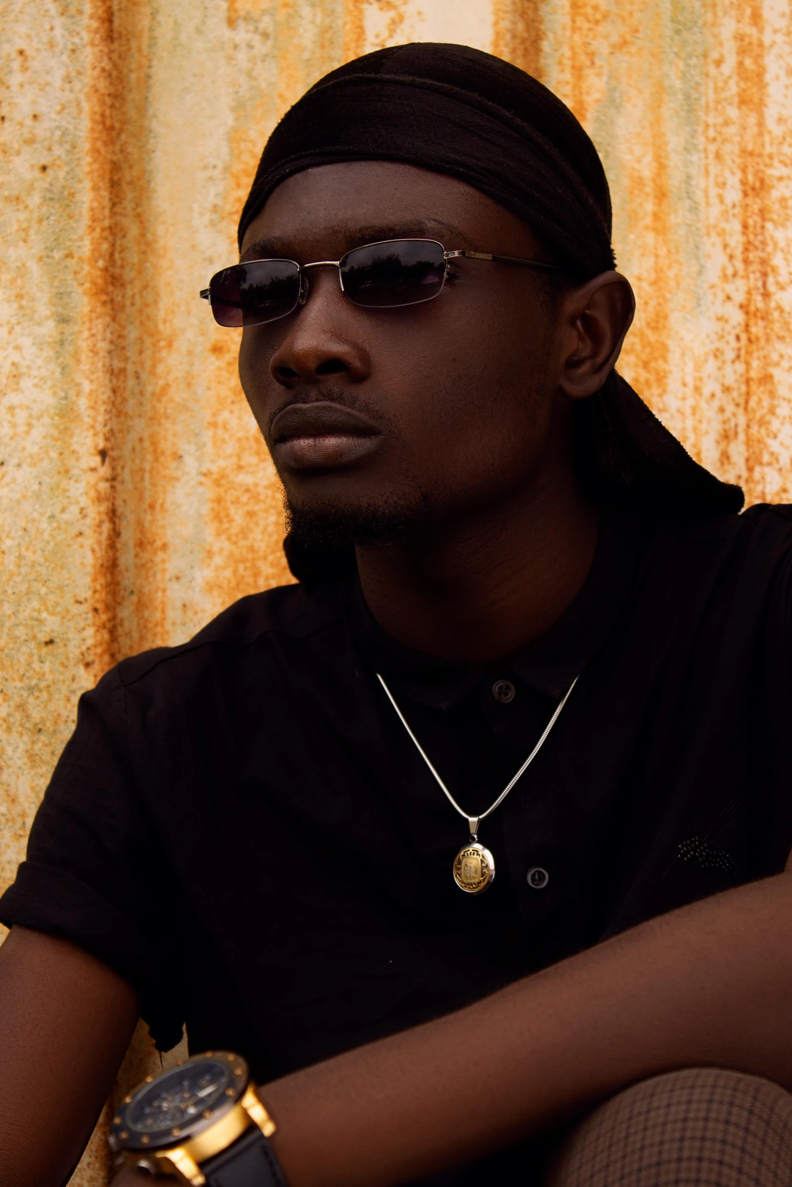 a man sitting in front of a rusty wall, an album cover, inspired by David Bailly, ( ( dark skin ) ), wearing shades, solarised, profile image