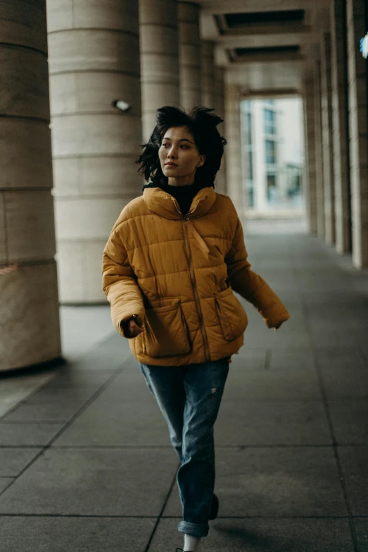 a woman in a yellow jacket walking down a sidewalk, pexels contest winner, puffer jacket, with two front pockets, running freely, muted colors. ue 5