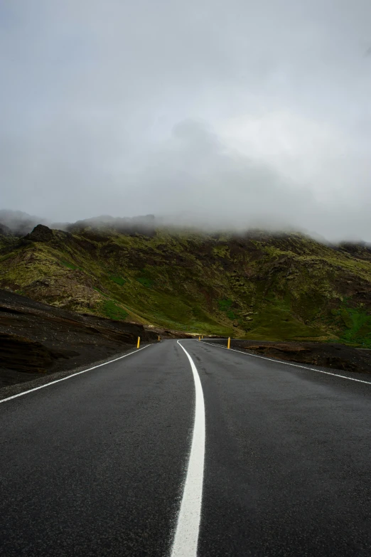 an empty road with a mountain in the background, by Hallsteinn Sigurðsson, pexels contest winner, under a gray foggy sky, some of the blacktop is showing, green hills, paved