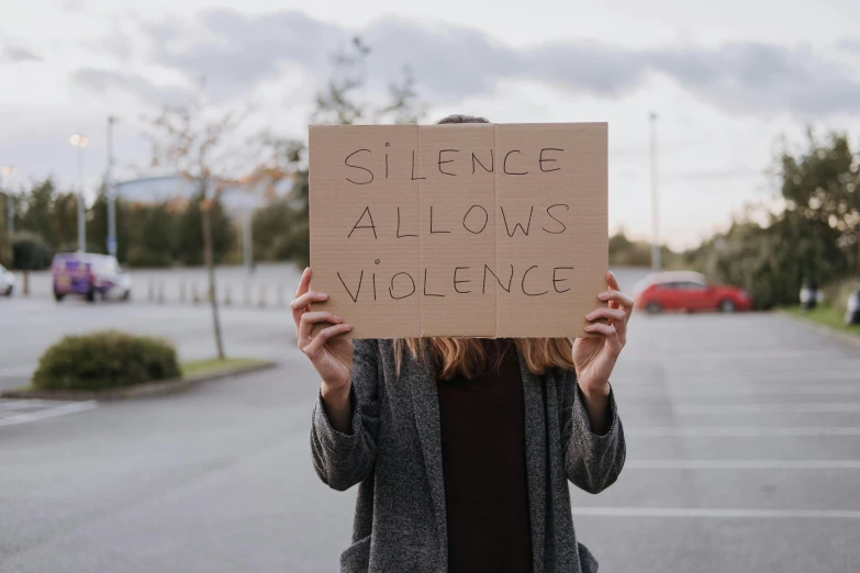 a woman holding a sign that says silence allows violence, a picture, by Emma Andijewska, shutterstock, instagram post, cardboard, gray, exterior shot