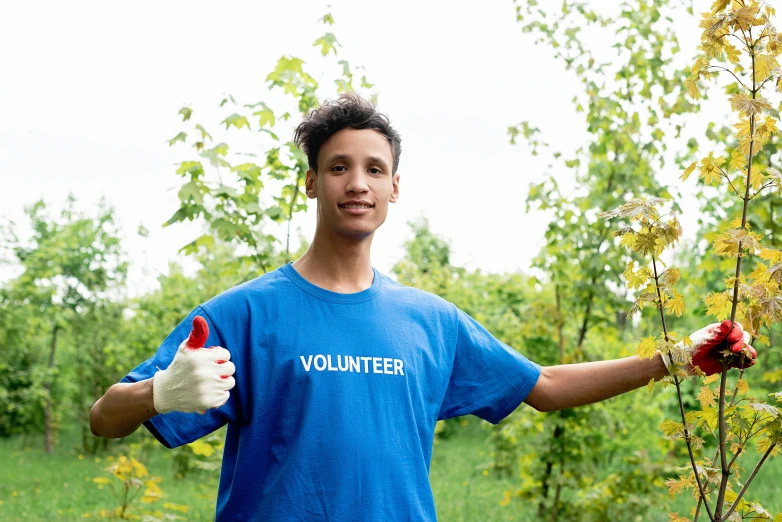a man in a blue shirt standing next to a tree, by Francis Helps, shutterstock, arbeitsrat für kunst, male teenager, giving a thumbs up to the camera, wearing gloves, promotional image