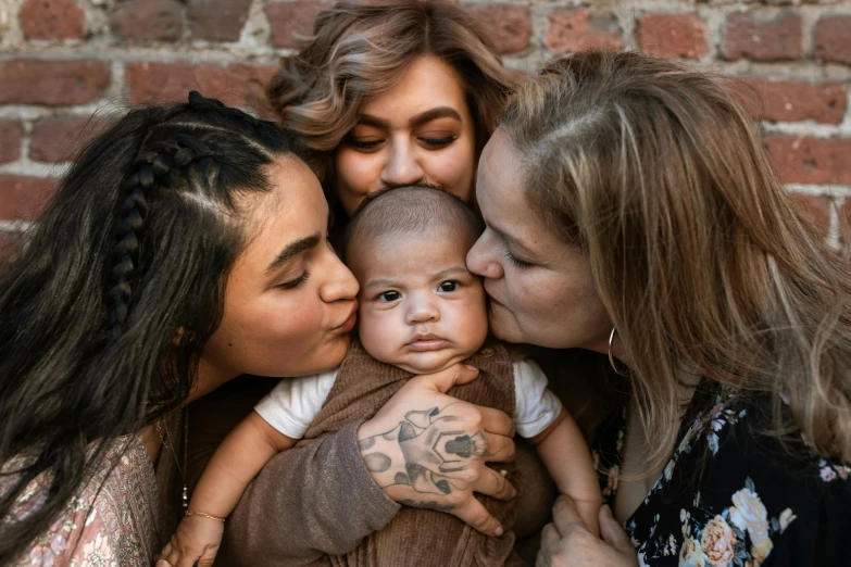 a group of women holding a baby in front of a brick wall, trending on pexels, lesbian embrace, close up portrait photo, tattooed, brown