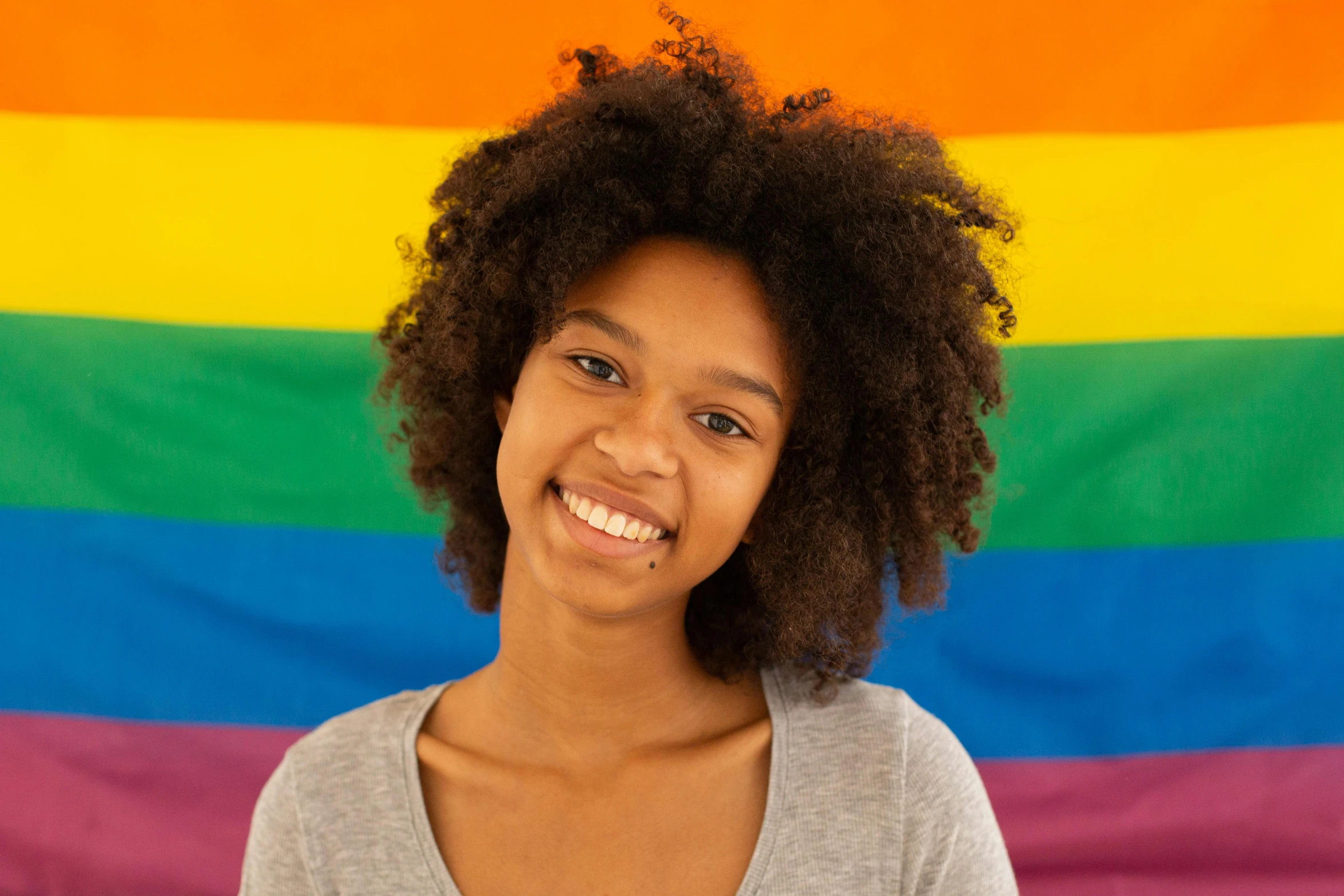 a woman standing in front of a rainbow flag, by Lily Delissa Joseph, trending on unsplash, dark short curly hair smiling, black teenage boy, her skin is light brown, official product photo