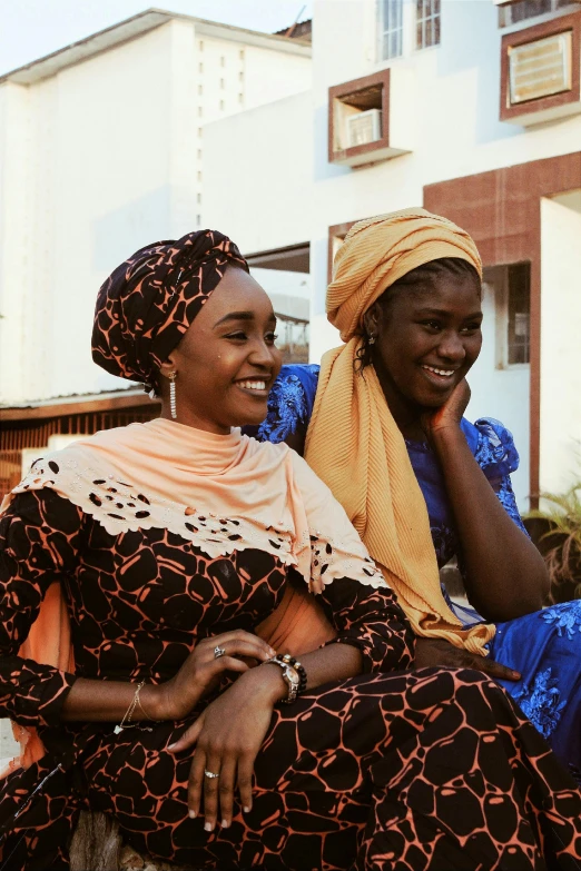 a couple of women sitting next to each other, hurufiyya, wearing an african dress, wearing a head scarf, smiling at each other, 2019 trending photo