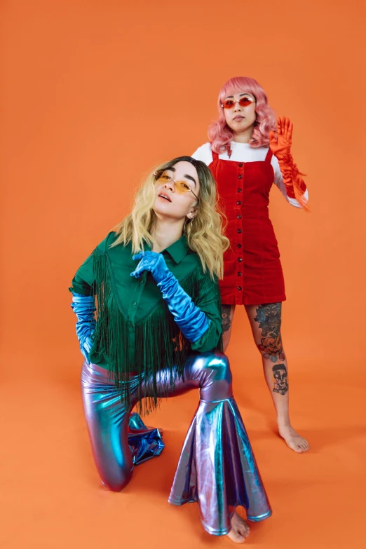 two women standing next to each other on an orange background, an album cover, trending on pexels, kitsch movement, glamourous cosplay, teal silver red, wearing festive clothing, bleached colours