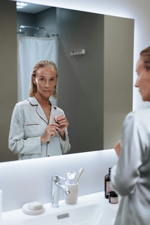 a woman standing in front of a bathroom mirror, happening, grey robes, facial precision, wearing pajamas, older woman