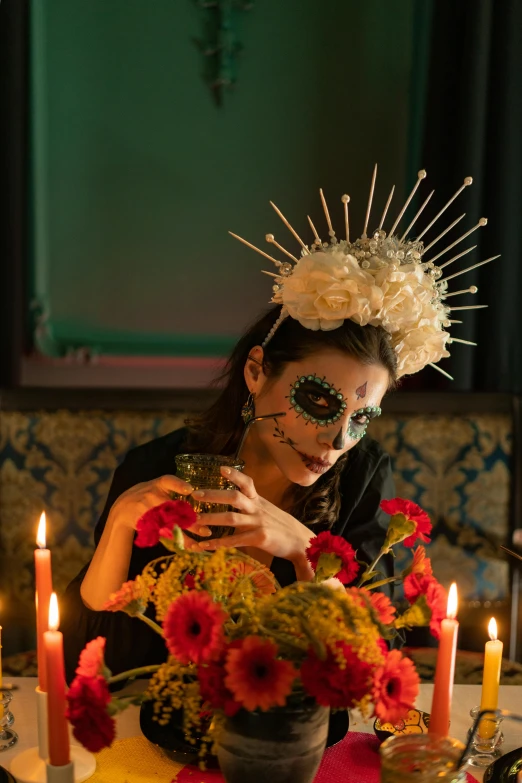 a woman sitting at a table with flowers and candles, inspired by Kahlo, vanitas, ram skull headpiece, holding a drink, holiday season, promotional shot