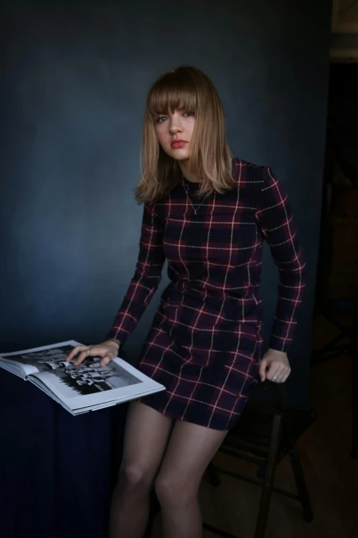 a woman standing next to a table holding a magazine, a colorized photo, inspired by Leila Faithfull, pexels contest winner, photorealism, portrait of taylor swift, plaid tights, studio portrait, center parted bangs