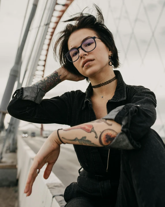 a woman sitting on the side of a bridge, a tattoo, trending on pexels, antipodeans, square rimmed glasses, non binary model, portrait image, arms folded