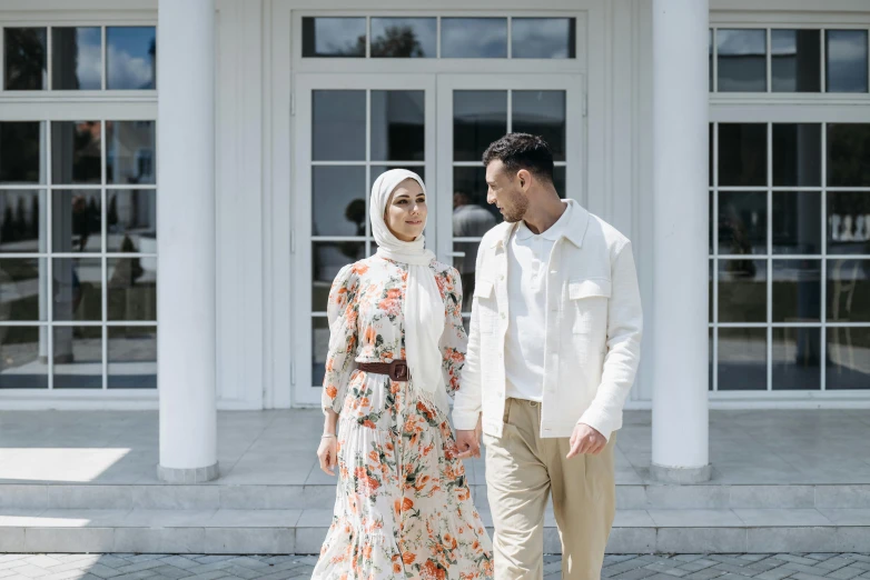 a man and a woman standing in front of a building, inspired by Nazmi Ziya Güran, pexels contest winner, hurufiyya, floral clothes, white hue, walking towards camera, a handsome