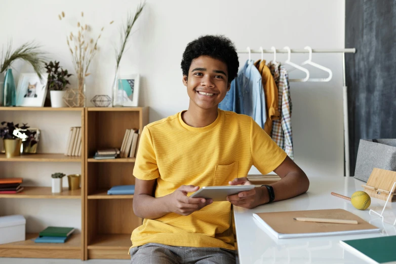 a young man sitting at a desk using a tablet computer, pexels contest winner, happening, yellow clothes, black teenage boy, posing for a picture, sitting on a store shelf