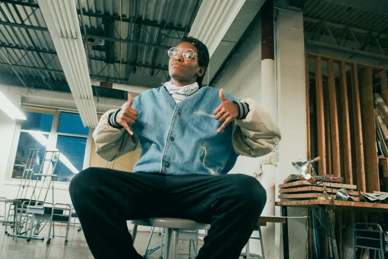 a man sitting on top of a chair in a room, visual art, young thug, stood in a lab, wearing small round glasses, sits on a finger