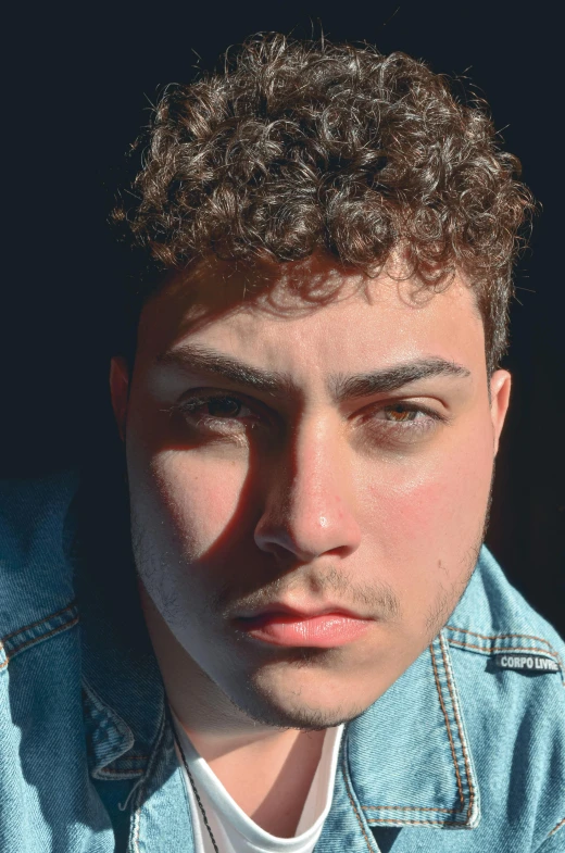 a close up of a person wearing a denim jacket, by Leo Michelson, big bold thick eyebrows, curls on top, asher duran, face is brightly lit