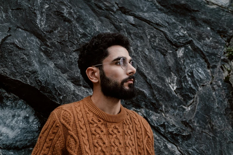 a man standing in front of a rock wall, a character portrait, trending on pexels, brown sweater, arab man light beard, man with glasses, androgynous person