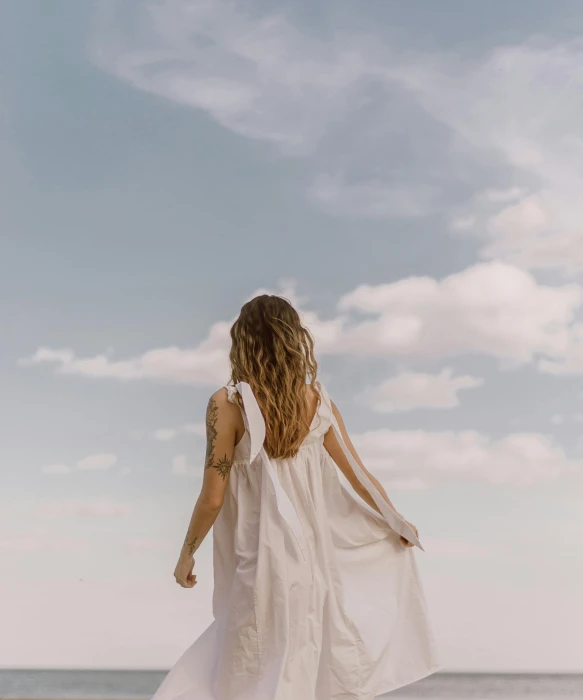 a woman in a white dress standing on a beach, trending on pexels, happening, flitting around in the sky, walking away, wearing a nightgown, partly cloudy