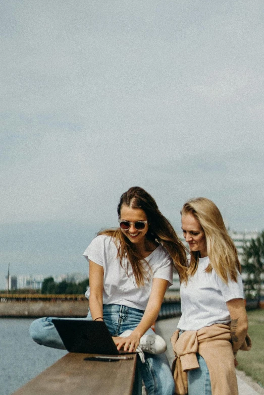 two women sitting on a bench looking at a laptop, by Niko Henrichon, trending on pexels, happening, wearing white v - neck top, on a riverbank, promotional image, happy friend