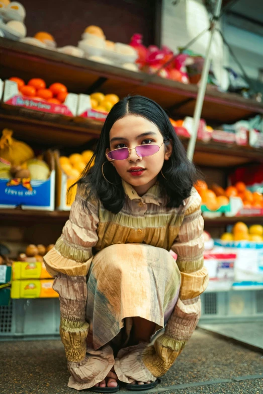 a woman sitting on the ground in front of a fruit stand, an album cover, trending on pexels, wavy long black hair and glasses, she looks like a mix of grimes, south east asian with round face, sitting on a store shelf