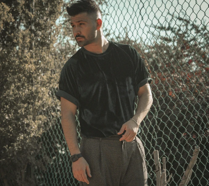 a man standing in front of a chain link fence, by Robbie Trevino, wearing pants and a t-shirt, with backdrop of natural light, zachary corzine, hollywood promotional image