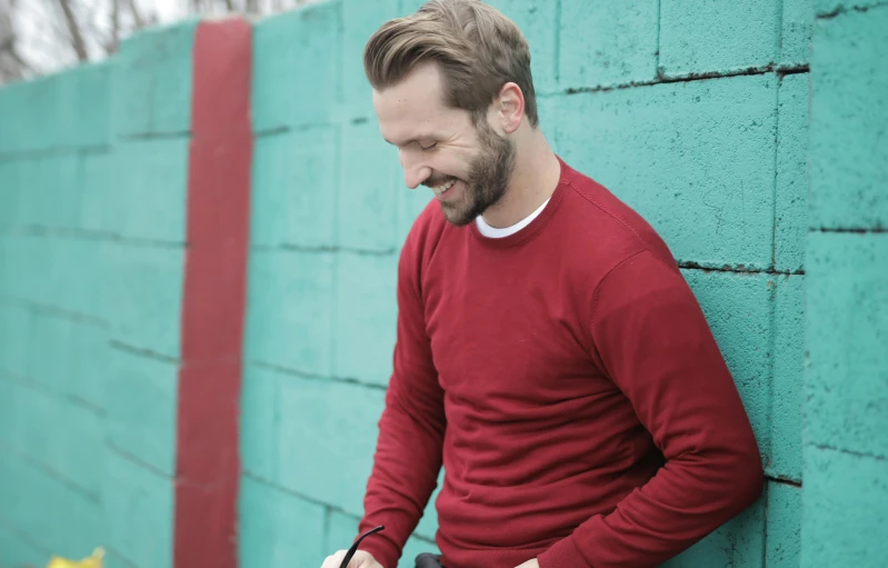 a man leaning against a wall using a cell phone, inspired by Alexander Robertson, pexels contest winner, happening, red sweater and gray pants, david beckham, receding hairline, short dark blond beard