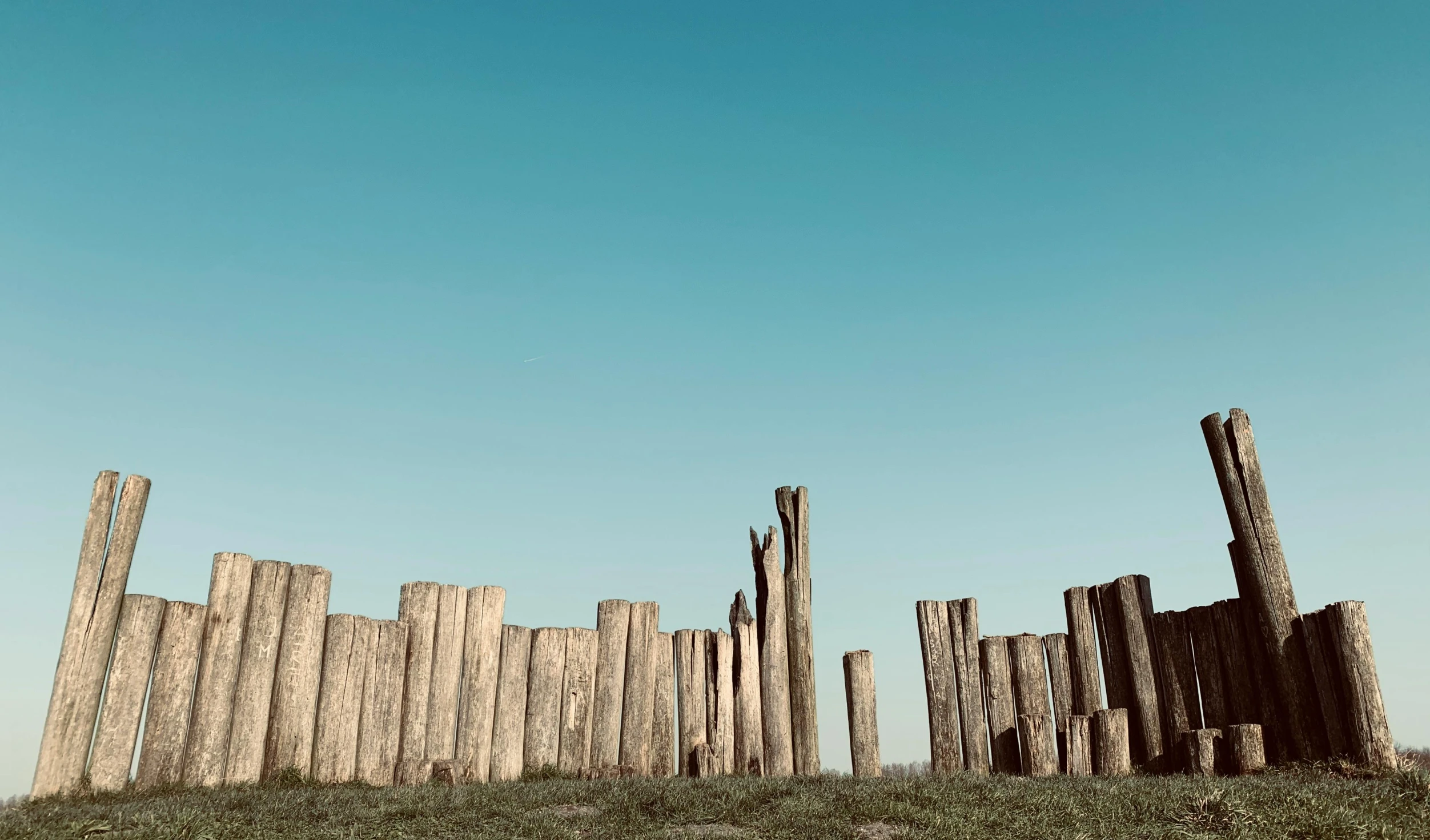 a wooden fence sitting on top of a lush green field, by Filip Hodas, land art, tall columns, driftwood, instagram photo, clear blue sky vintage style