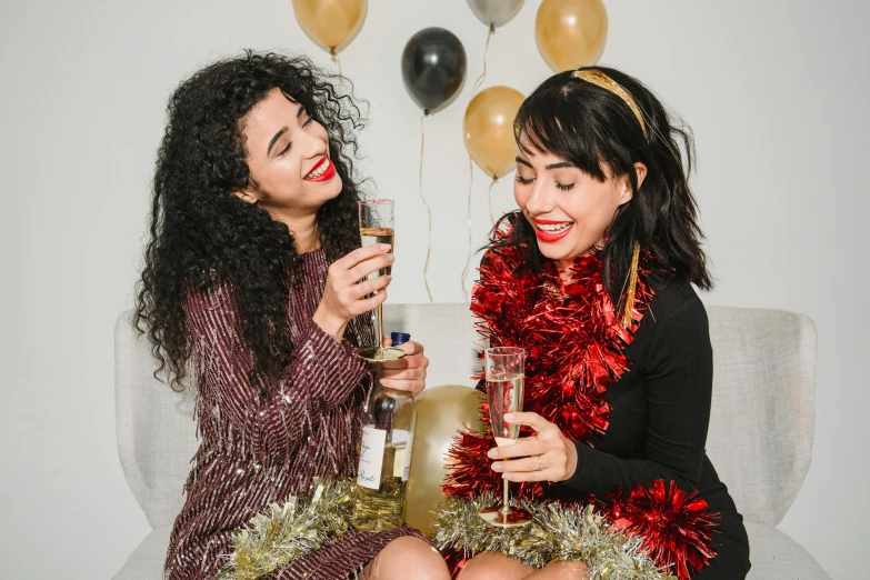 a couple of women sitting on top of a couch, pexels, happening, new years eve, drinking wine, gold and red accents, with curly black and silver hair