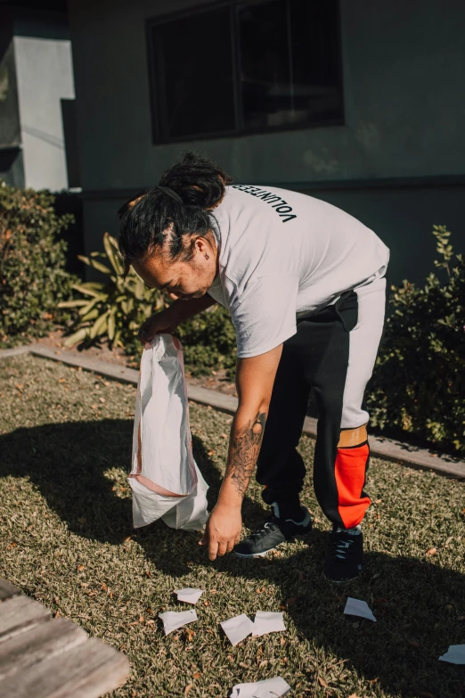 a man that is standing in the grass with a bag, federation clothing, pulling weeds out frantically, mullet, in a suburb