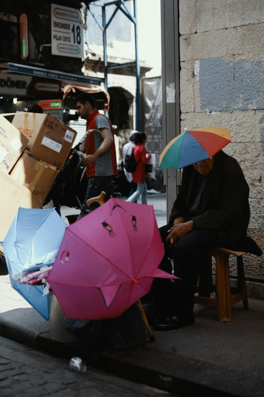 a group of people sitting on a sidewalk with umbrellas, by Nathalie Rattner, jerusalem, paul barson, a still of kowloon, cardboard