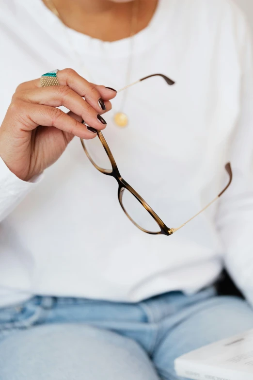 a woman sitting on a couch holding a pair of glasses, by Jessie Algie, minimalism, neck zoomed in, thin gold details, wearing a white sweater, zoomed in