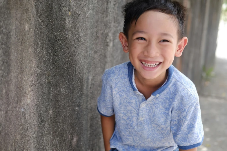 a young boy smiles as he leans against a wall, inspired by Reuben Tam, happily smiling at the camera, avatar image, commercial shot, caring fatherly wide forehead