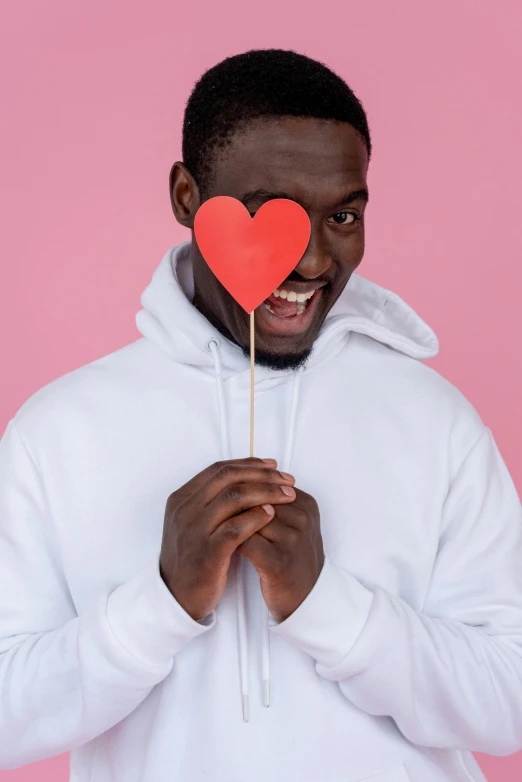 a man in a white hoodie holding a red heart on a stick, trending on pexels, brown skin man with a giant grin, dating app icon, pink, cardboard cutout