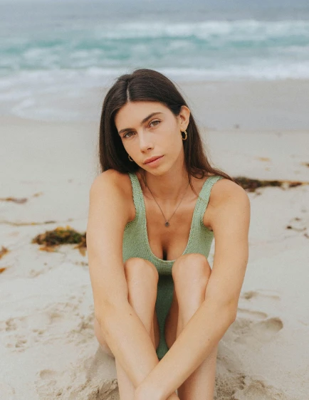 a woman sitting on top of a sandy beach, an album cover, inspired by Elsa Bleda, trending on pexels, renaissance, green swimsuit, nonbinary model, madison beer girl portrait, looking directly at the camera