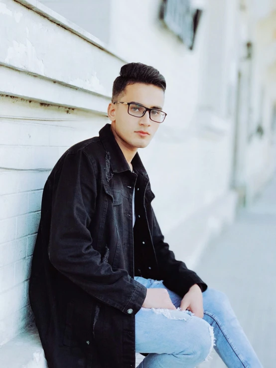 a young man sitting on the side of a building, by Robbie Trevino, wearing black rimmed glasses, dasha taran, profile image, androgynous person